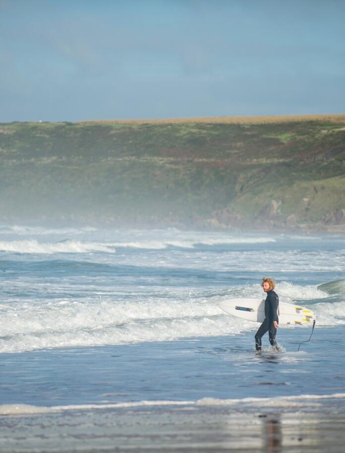 Surfer standing in teh blue shallow waters of teh sandy beach with waves crashing 