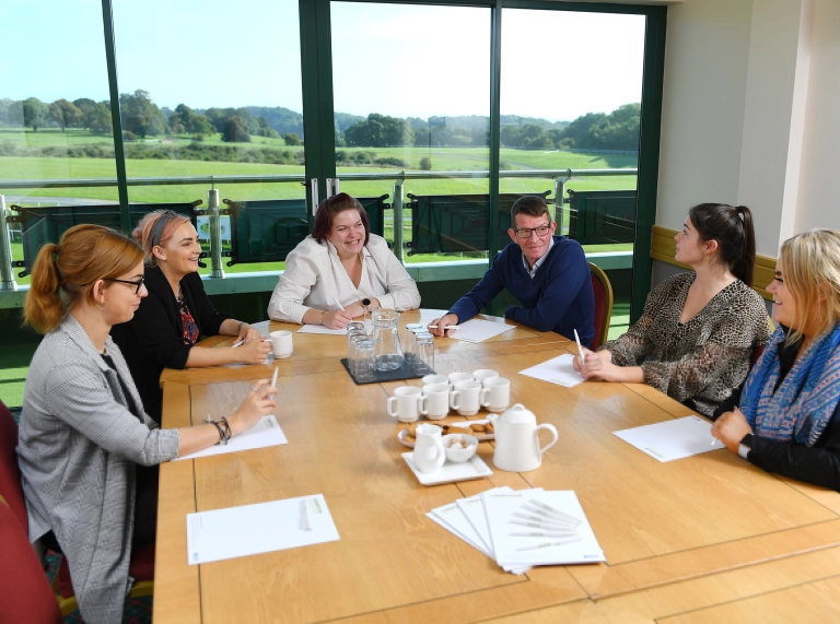 A group of people sat around a table having a meeting with tea and biscuits in the centre.