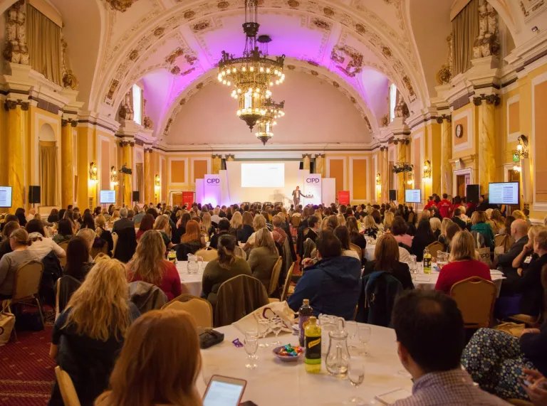 People sat around tables looking toward a speaker and screen in a grand hall.