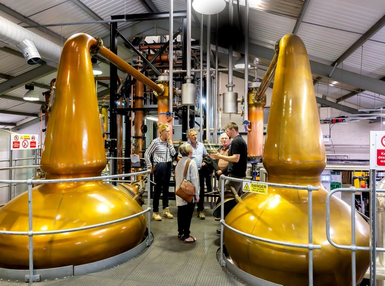 A group of people taking a tour of a distillery, next to two golden vats.