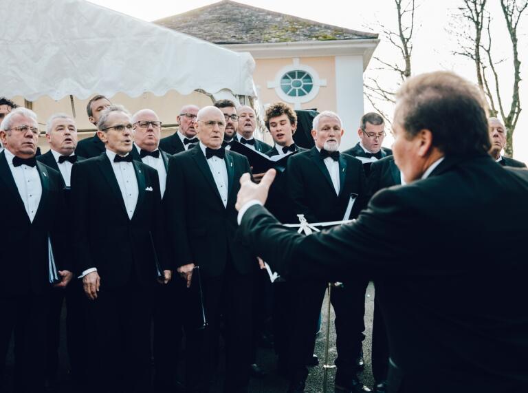 Male choir dressed in black suits with white shirts signing as conductor instructs them