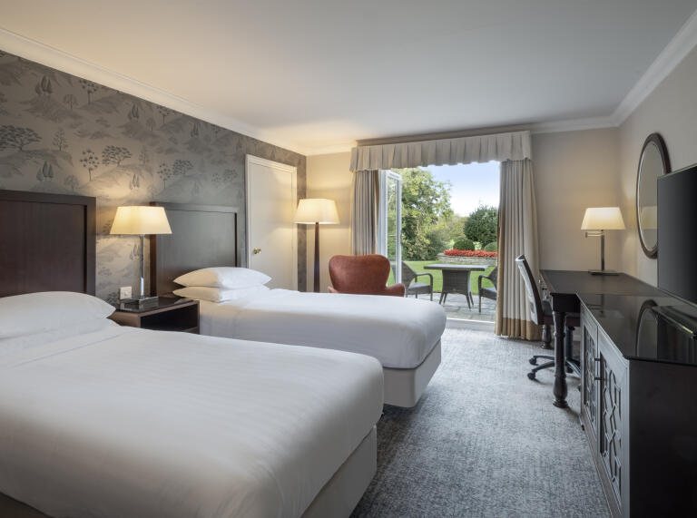 twin beds with white sheets and  adesk and mirro with chair overlooking the patio doors onto the green grass and surrounding trees