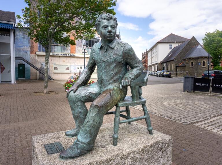 stone statue of Dylan Thomas sitting  on stone with gardens behind