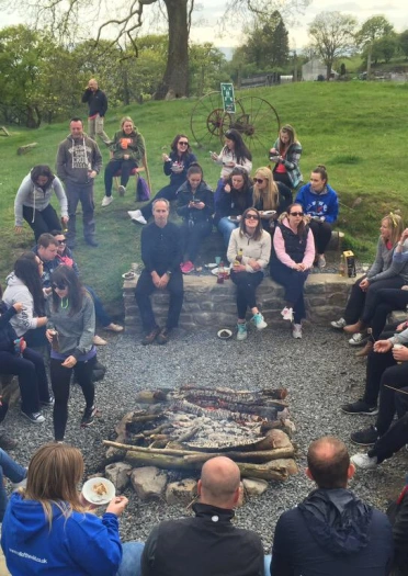 A group of people sitting around a campfire eating a barbecue.