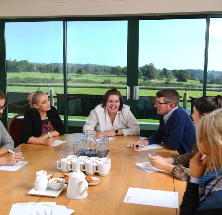 People around a meeting table with views of a racecourse beyond.