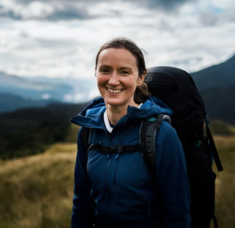 Claire Copeman smiling at the camera wearing outdoor gear and carrying a backpack with mountains in the background.