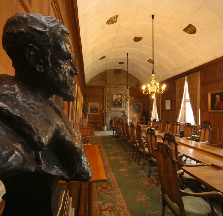 The council chamber witha   head statue of a man low chandelier lights over a log table with fireplace at the end.