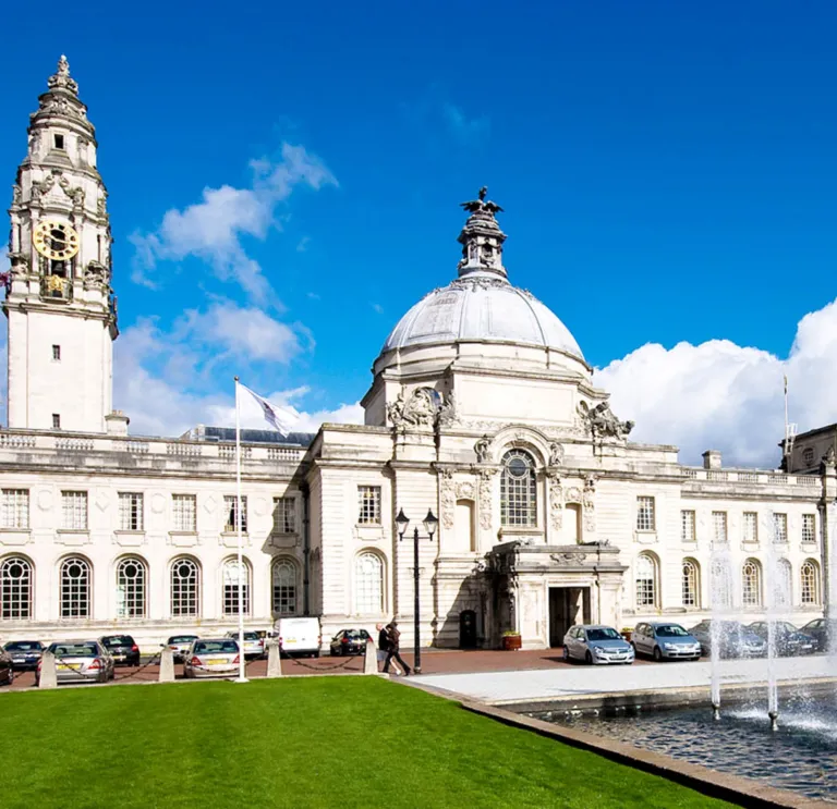 External of building white brick glass windows and clock on tower, with fountain and lawn green in the forefront.