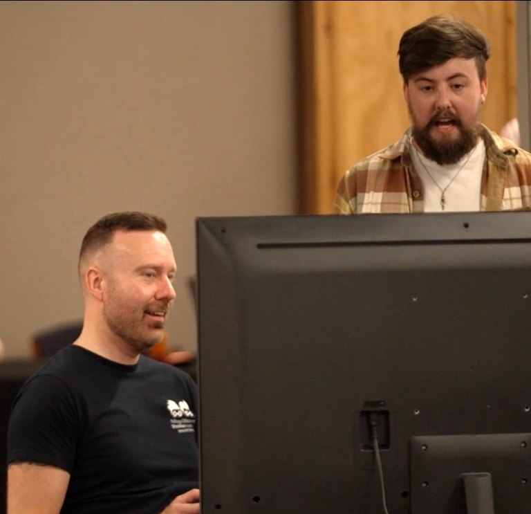 Ambassador Richard Hebblewhte sitting at a computer screen talking with a collegue standing behind him in a checked shirt 