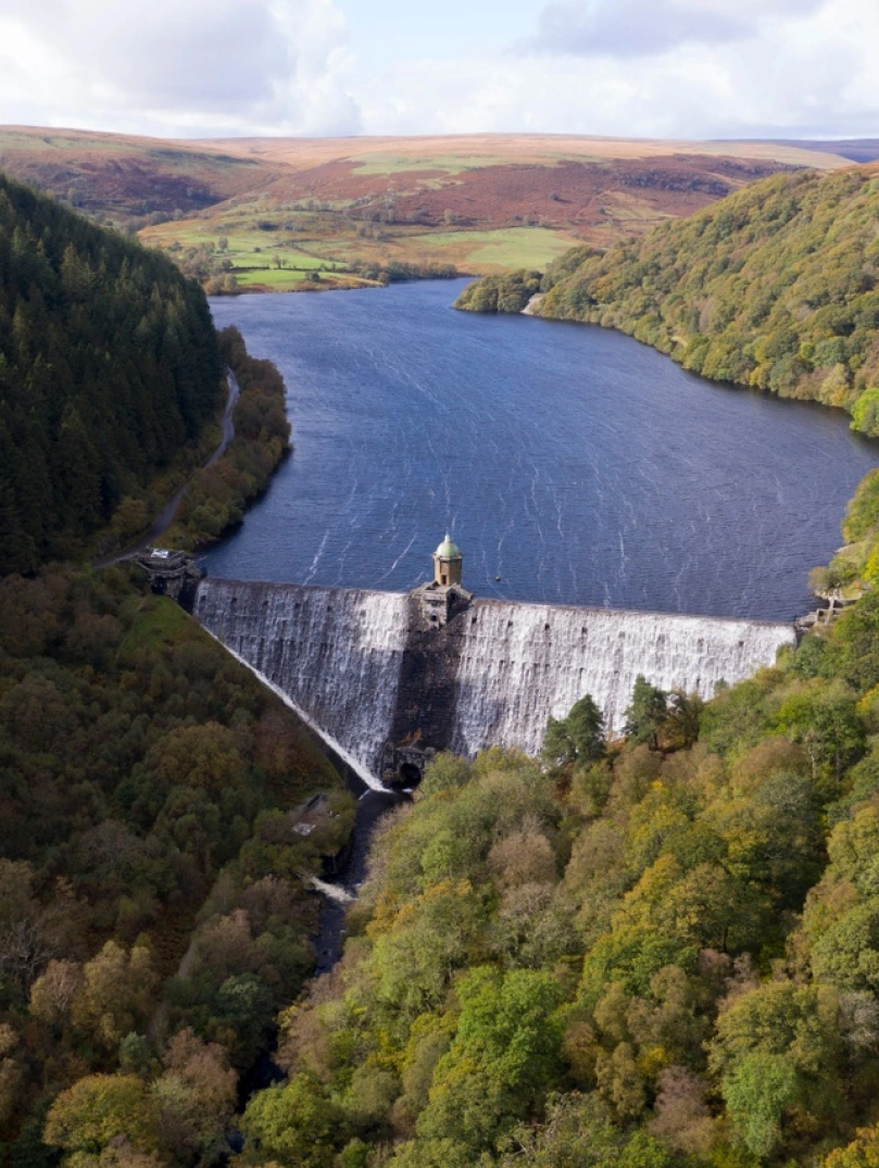 Aerial view of a river flowing over an open dam surrounded by trees and mountains.
