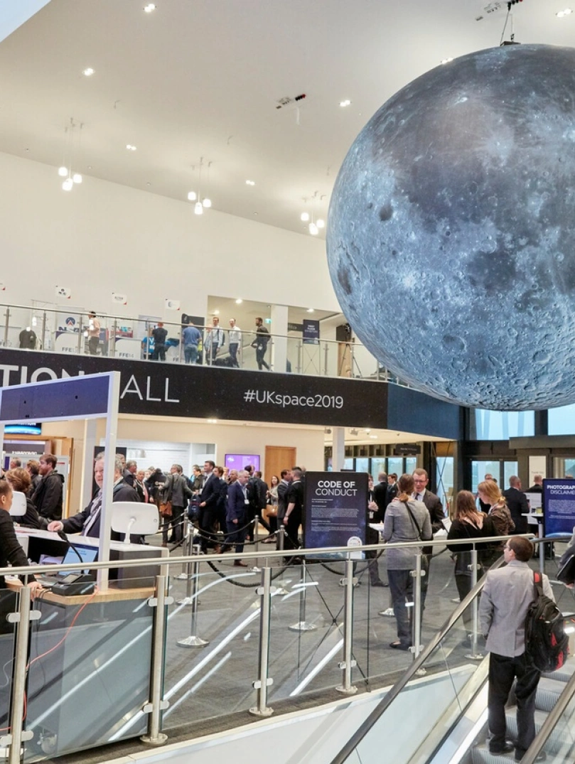 Exhibition hall with people coming up esculators to reception desks and a large grey space globe suspended from the ceiling.