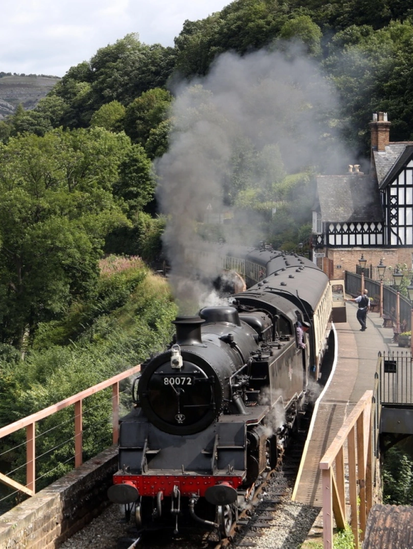 A steam train pulling out of a station alongside a lake.