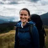 Claire Copeman smiling at the camera wearing outdoor gear and carrying a backpack with mountains in the background.